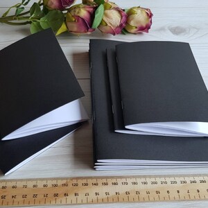May include: A stack of three black notebooks with white pages. The notebooks are closed and have a smooth, matte finish.