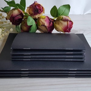 May include: A stack of black notebooks with a simple design. The notebooks are bound with a stitched spine and have a smooth, matte finish.