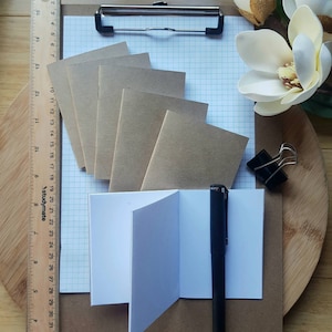 May include: A stationery flat lay featuring a clipboard with graph paper, several small brown notebooks, and an open white notebook. A black pen and a ruler are also present. A white flower and a black binder clip complete the arrangement.