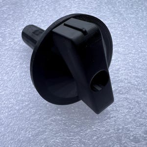 May include: A black plastic bottle stopper with a round base and a rectangular top. The stopper has a cylindrical stem and a hole on the side. The object is on a textured, light-colored surface.