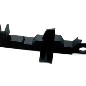 Puede incluir: Un componente de plástico negro con una forma única, que presenta una base rectangular, múltiples secciones elevadas y un bucle circular en un extremo. El diseño incluye una aleta vertical y una sección con muescas. Sobre fondo blanco.