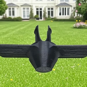 May include: A black lawn aerator tool with a wing-like design. The tool has two vertical prongs and horizontal wings, designed to penetrate the soil. The tool is resting on a green lawn with a large house in the background.