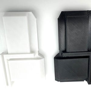 May include: Two 3D-printed objects, one white and one black, are displayed. Each has a rectangular shape with a stepped design. The objects appear to be small, possibly components or decorative items. The white object is on the left, and the black object is on the right.