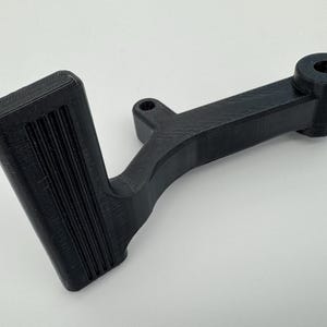 May include: A black, 3D-printed automotive component, possibly a pedal or lever. The part has a textured, rectangular footpad with vertical grooves and a curved arm. It features a cylindrical end with a hole.