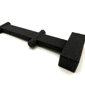 May include: A black, textured, T-shaped object with a long, narrow central bar, a cylindrical protrusion on one side, and a rectangular block at the end. The surface has a rough, granular finish throughout, and the structure is rigid and uniform.