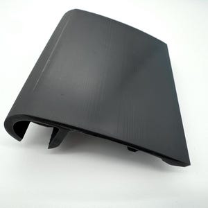 May include: A black plastic automotive part with a textured surface. The part has a curved edge and a rectangular shape with a slight angle. The part is likely a trim piece or cover for a vehicle.