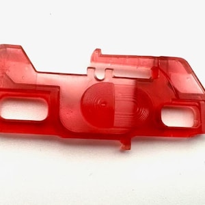 Puede incluir: Un componente de plástico rojo translúcido con una forma única. Presenta un cuerpo rectangular con esquinas redondeadas, dos recortes rectangulares y un diseño circular en el centro. El objeto está sobre un fondo blanco.