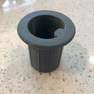 Puede incluir: Un adaptador de plástico gris con una abertura circular y una muesca en el lateral. El adaptador tiene un diseño acanalado en la parte inferior.