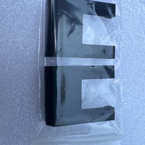 May include: Two black, rectangular, U-shaped components are sealed in a clear plastic bag. The components have a glossy finish and appear to be made of a solid material. The bag is sealed with a zipper closure.