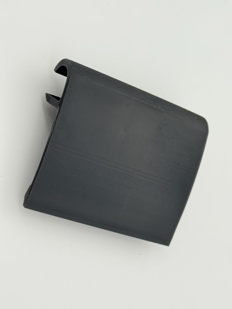Pode incluir: Um componente de pl&aacute;stico preto, retangular, com uma borda curva e uma superf&iacute;cie ranhurada. O componente tem uma pequena aba saliente em um lado. O objeto est&aacute; sobre um fundo branco.