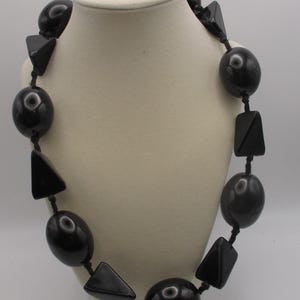 May include: A necklace featuring alternating black beads in spherical and triangular shapes strung on a black cord. The smooth, glossy black components are arranged in a repeating pattern around the length of the jewelry piece.