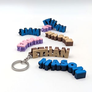 Op de afbeelding: Een verzameling kleurrijke 3D-geprinte naam sleutelhangers. De namen, waaronder Liam, Emma, Ethan en Mason, zijn in verschillende kleurencombinaties zoals blauw, roze en bruin. Eén sleutelhanger heeft een zilveren ring.