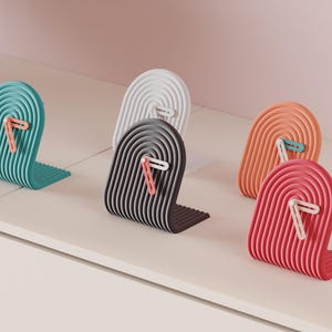 Op de afbeelding: Een verzameling kleurrijke boekensteunen in tinten van turquoise, wit, grijs, oranje en rood. Elke boekensteun heeft een gebogen, geribbeld ontwerp en een contrasterend paperclip accent. De boekensteunen staan op een wit oppervlak.