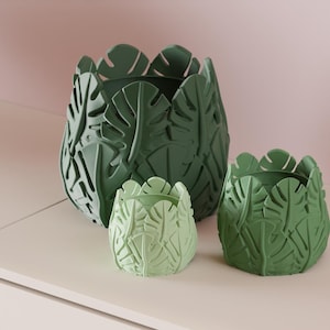Op de afbeelding: Drie decoratieve plantenpotten in verschillende maten en groentinten. De potten hebben de vorm van bladeren en staan op een wit oppervlak. De grootste pot is donkergroen, de kleinste lichtgroen. Afmetingen in centimeters.