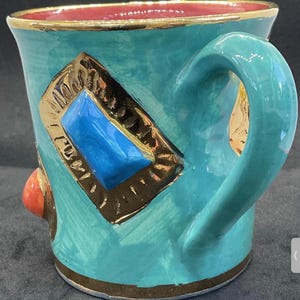 May include: A turquoise ceramic mug with a gold rim and handle. The mug features a gold-framed diamond shape with a blue centre. An orange accent is on the side, and the interior is red.