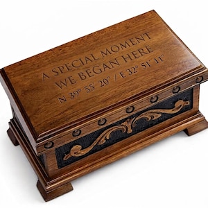 Peut inclure: Une boîte rectangulaire en bois avec une finition brun foncé, portant l'inscription gravée A SPECIAL MOMENT WE BEGAN HERE N 39° 55' 20" / E 32° 51' 11" sur le couvercle. Le panneau avant présente un ornement sculpté en bois sur un fond noir texturé.