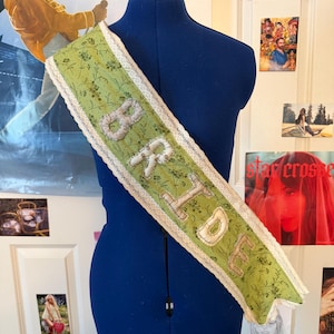 May include: A sage green sash featuring a delicate dark green floral pattern and white lace trim along the edges. The word BRIDE is appliqued in white fabric letters across the center of the sash, which is displayed on a blue dress form.