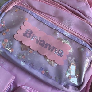 Puede incluir: Una mochila rosa y morada con múltiples compartimentos con cremallera. La mochila presenta un bolsillo transparente con elementos decorativos y una etiqueta con el nombre rosa que dice "Brianna" en letras brillantes. La bolsa está adornada con lentejuelas y otros adornos.