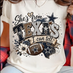 May include: A cream-colored t-shirt featuring a graphic print with the text "She's from TEXAS I can tell". The design includes a football, a star, a flower, and a map of Texas, decorated with leopard print, glitter, and navy blue accents.