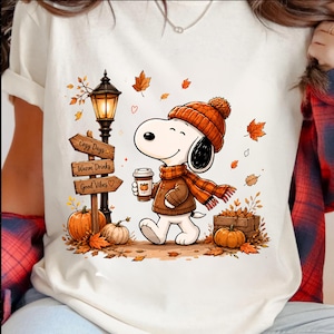 Op de afbeelding: Een crèmekleurig t-shirt met een afbeelding van een stripfiguurhond met een oranje gebreide muts en sjaal, die een koffiebeker vasthoudt. De scène bevat een lantaarnpaal, houten borden met Cozy Days, Warm Drinks, Good Vibes en pompoenen.