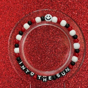 May include: A beaded bracelet with red, white, black, and smiley face beads. The bracelet is displayed on a clear, circular stand. The words "INTO THE SUN" are spelled out in black letter beads around the stand.