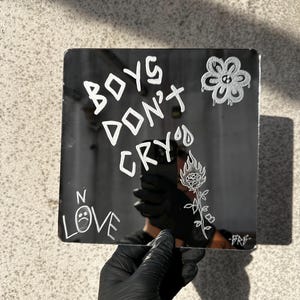 Può includere: Uno specchio nero con scritta bianca che dice "Boys Don't Cry" e "N Love" con un'emoji di faccia triste. C'è un fiore bianco e una rosa bianca con fiamme sullo specchio.