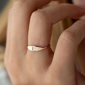 May include: A rose gold ring with a rectangular face, featuring the letter "E" engraved on it. The ring is worn on a finger, showcasing its delicate design and minimalist style. The ring is a piece of jewelry.