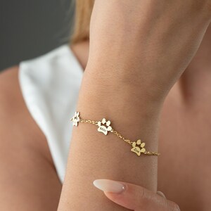 May include: Gold-coloured bracelet with three paw-shaped charms. Each charm is engraved with a name. The bracelet is worn on a wrist, against a soft grey background.