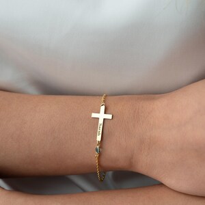 May include: A gold-colored bracelet featuring a cross pendant. The cross has a vertical bar with the name "Sarah" engraved on it. The bracelet includes a small, light blue gemstone and is worn on a wrist.