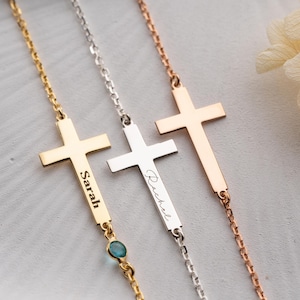 May include: Three personalized cross necklaces in gold, silver, and rose gold. Each necklace features a cross pendant with a name engraved on a vertical bar. The gold necklace has a small blue gemstone. The names "Sarah" and "Rachel" are visible.