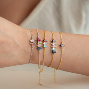 May include: Several fine chain bracelets, each adorned with marquise-cut gemstones in various colours. The bracelets are silver and gold, with stones in red, pink, blue, and clear. The bracelets are displayed on a wrist.