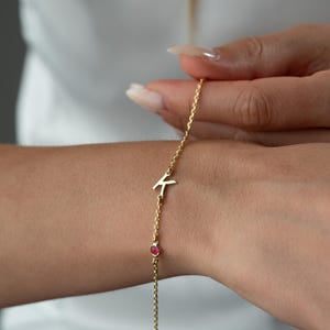 May include: A delicate gold bracelet with the letter "K" and a small, round, red gemstone. The bracelet is held on a wrist, highlighting its fine chain and minimalist design. The jewellery is a stylish accessory.
