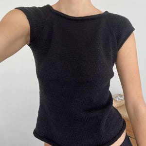 May include: A black knit top with short sleeves. The top has a scoop neckline and a fitted silhouette. The fabric appears to be a textured knit. The top is a solid black color.