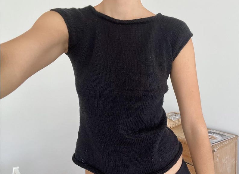 May include: A black knit top with short sleeves. The top has a scoop neckline and a fitted silhouette. The fabric appears to be a textured knit. The top is a solid black color.