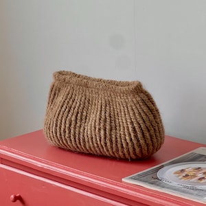 Puede incluir: Un bolso de punto de color marrón con una textura acanalada y una forma redondeada y suave. El bolso presenta una abertura superior fruncida y una construcción gruesa similar a la lana, con un tono tierra neutro y sólido en todo el exterior.