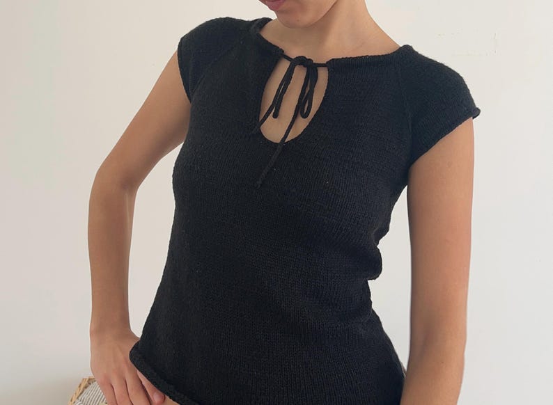 May include: Black knit top with a keyhole neckline and short sleeves. The top features a tie detail at the neckline and a textured knit pattern. The garment is a solid black color.