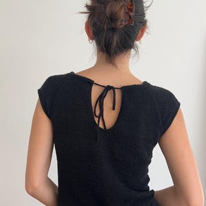 May include: Black knit top with a keyhole back and tie closure. The short-sleeved top has a scoop neckline and a ribbed hem. The model's hair is pulled back with a brown hair clip. The top is made of a textured knit fabric.