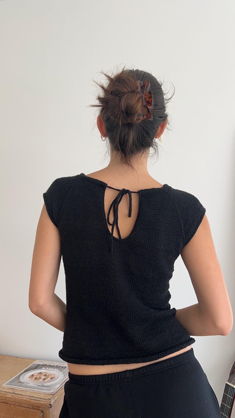 May include: Black knit top with a keyhole back and tie closure. The short-sleeved top has a scoop neckline and a ribbed hem. The model's hair is pulled back with a brown hair clip. The top is made of a textured knit fabric.
