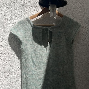 May include: A light green knitted top with short sleeves and a keyhole back detail. The top is displayed on a wooden hanger against a textured white wall. A black light fixture is above the hanger.