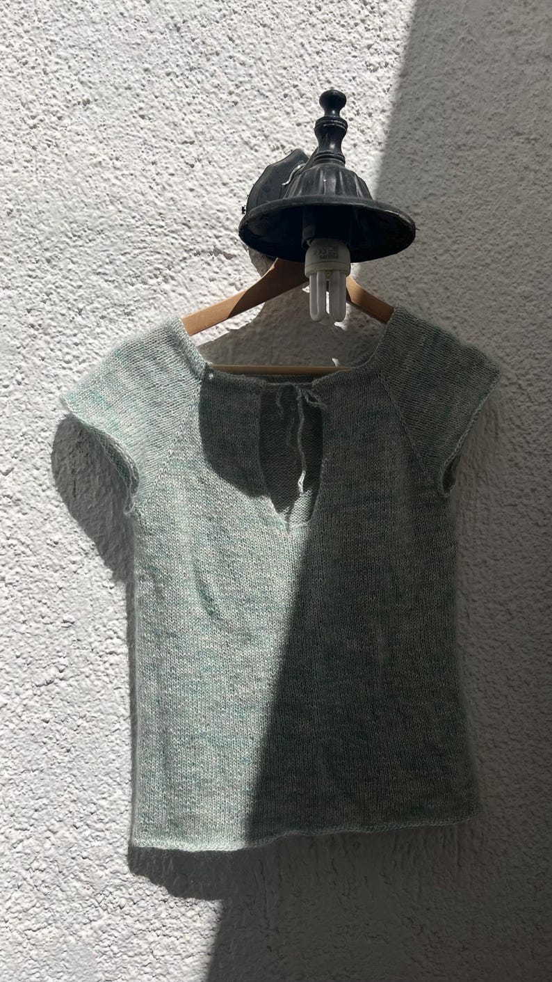 May include: A light green knitted top with short sleeves and a keyhole back detail. The top is displayed on a wooden hanger against a textured white wall. A black light fixture is above the hanger.