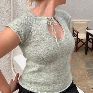 May include: A light green knitted top with short sleeves and a keyhole neckline. The top has a tie detail at the neckline and a slightly textured appearance. The garment is worn with a black skirt.