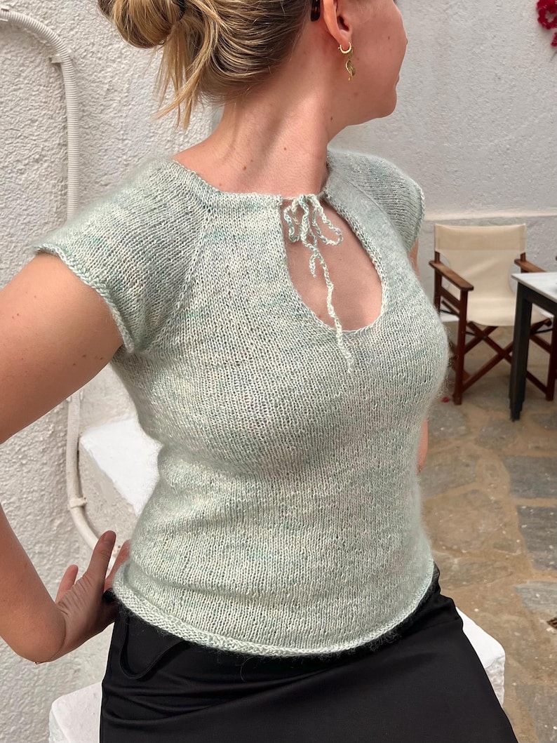 May include: A light green knitted top with short sleeves and a keyhole neckline. The top has a tie detail at the neckline and a slightly textured appearance. The garment is worn with a black skirt.