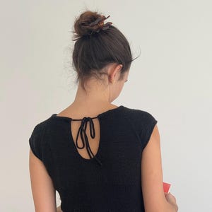 May include: A black knit top with a keyhole back and tie detail. The top has short sleeves and a textured appearance. The person is wearing a hair clip and earrings. The background is a plain white wall.