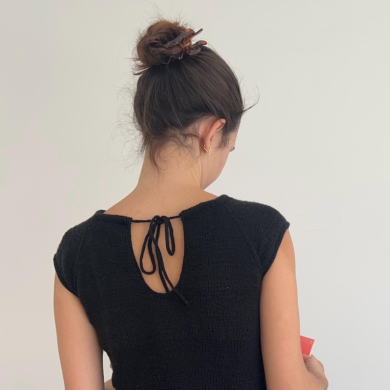 May include: A black knit top with a keyhole back and tie detail. The top has short sleeves and a textured appearance. The person is wearing a hair clip and earrings. The background is a plain white wall.