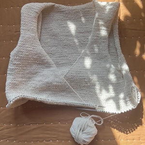 May include: A cream-colored, hand-knitted vest with a V-neck design. The vest is in progress, with knitting needles and a ball of white yarn visible. The background is a brown surface with sunlight and shadows.