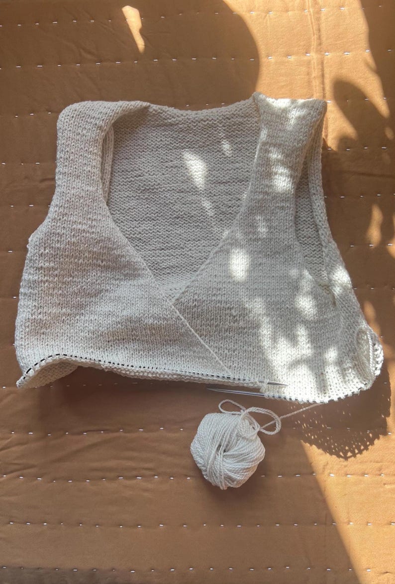 May include: A cream-colored, hand-knitted vest with a V-neck design. The vest is in progress, with knitting needles and a ball of white yarn visible. The background is a brown surface with sunlight and shadows.