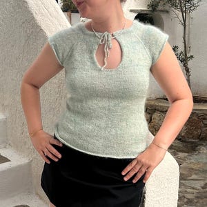 May include: A light green knitted top with a keyhole neckline and short sleeves. The top is made of a soft, textured yarn and has a tie detail at the neckline. The model is wearing a black skirt.