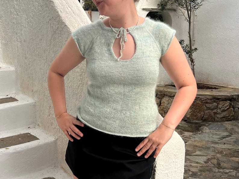 May include: A light green knitted top with a keyhole neckline and short sleeves. The top is made of a soft, textured yarn and has a tie detail at the neckline. The model is wearing a black skirt.