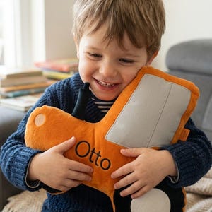 Könnte beinhalten: Ein Plüschtraktor-Spielzeug in Orange, Grau und Schwarz mit einem schwarzen Auspuffrohr. Auf der Seite des Traktors ist der Name Otto in schwarzem Garn eingestickt. Das Spielzeug hat eine weiche, strukturierte Oberfläche und ein großes schwarzes Rad.