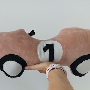 May include: A plush toy shaped like a vintage race car in a dusty pink color. The car features a white circular patch on the side with a black number one, black and white wheels, and four black stitched lines on the front hood area.